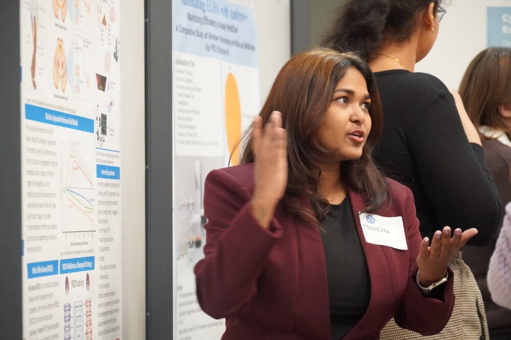 A woman wearing a maroon blazer and name tag labeled "Moulika" is presenting a scientific poster at a conference or academic event. She is speaking animatedly and using hand gestures to explain her work to others. with scientific posters in the background
